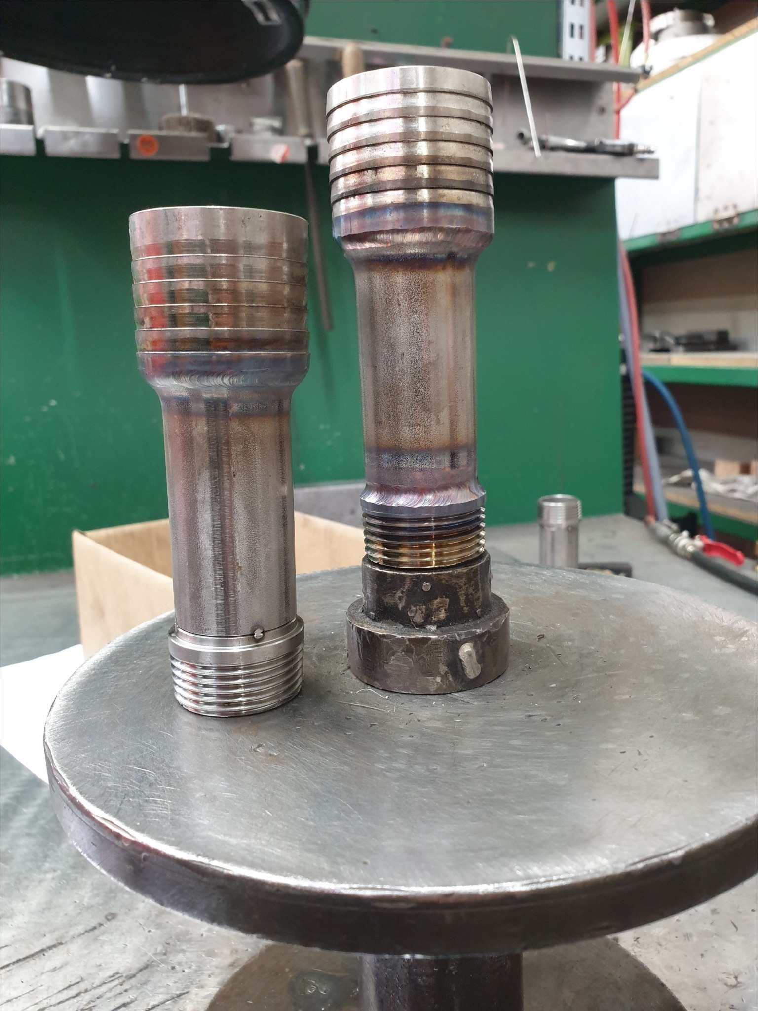 Dwa metalowe elementy z widocznymi śladami obróbki, w tym spoiny, umieszczone na metalowym stole warsztatowym w hali produkcyjnej.