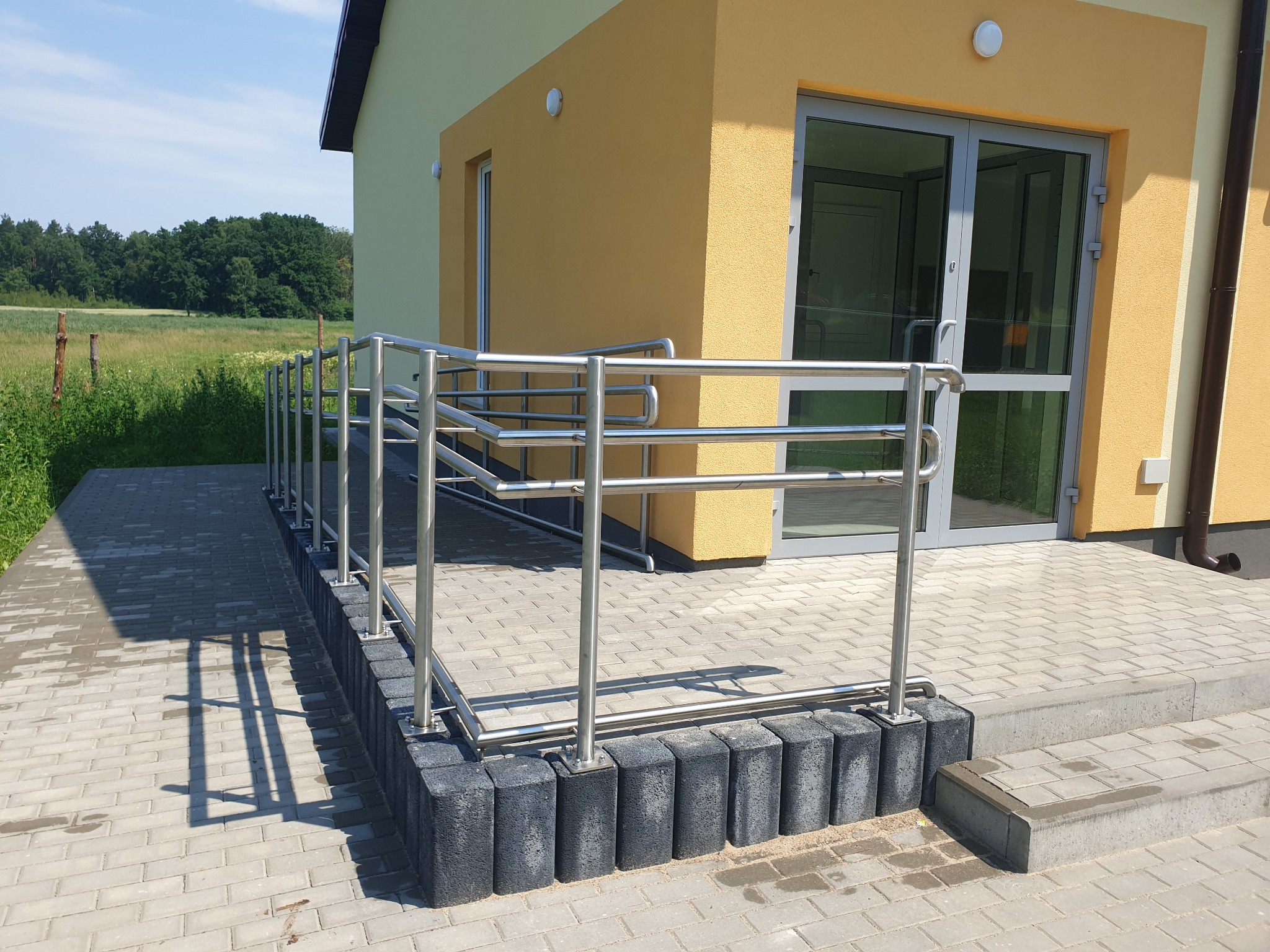 Zewnętrzna, trzypoziomowa, nierdzewna konstrukcja zabezpieczająca schody i podjazd dla niepełnosprawnych przy budynku w Olsztynie, zamontowana na betonowej kostce.