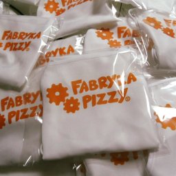 Sylwia Cywicka Składzik Prezentów - Stos zapakowanych w folię białych fartuchów z pomarańczowym nadrukiem 'Fabryka Pizzy', widoczne detale szwów i zagięć materiału.