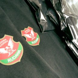 Sylwia Cywicka Składzik Prezentów - Ciemnoszara bluza dresowa z naszytymi herbami Akademii Piłkarskiej Rudawa, częściowo zapakowana w foliowe opakowanie.