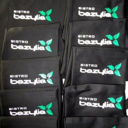 Sylwia Cywicka Składzik Prezentów - Partia czarnych fartuchów roboczych z nadrukowanym logo 'Bistro Bazylia' w kolorze białym i zielonym.