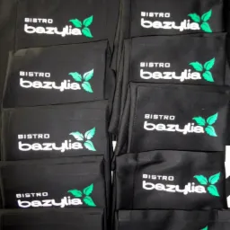 Partia czarnych fartuchów roboczych z nadrukowanym logo 'Bistro Bazylia' w kolorze białym i zielonym.