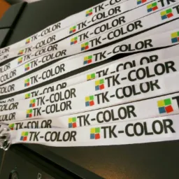 Partia białych smyczy z nadrukowanym czarnym napisem 'TK-COLOR' i kolorowym logo z czterech kwadratów (czerwony, żółty, zielony, niebieski), leżących na czarnej drukarce.
