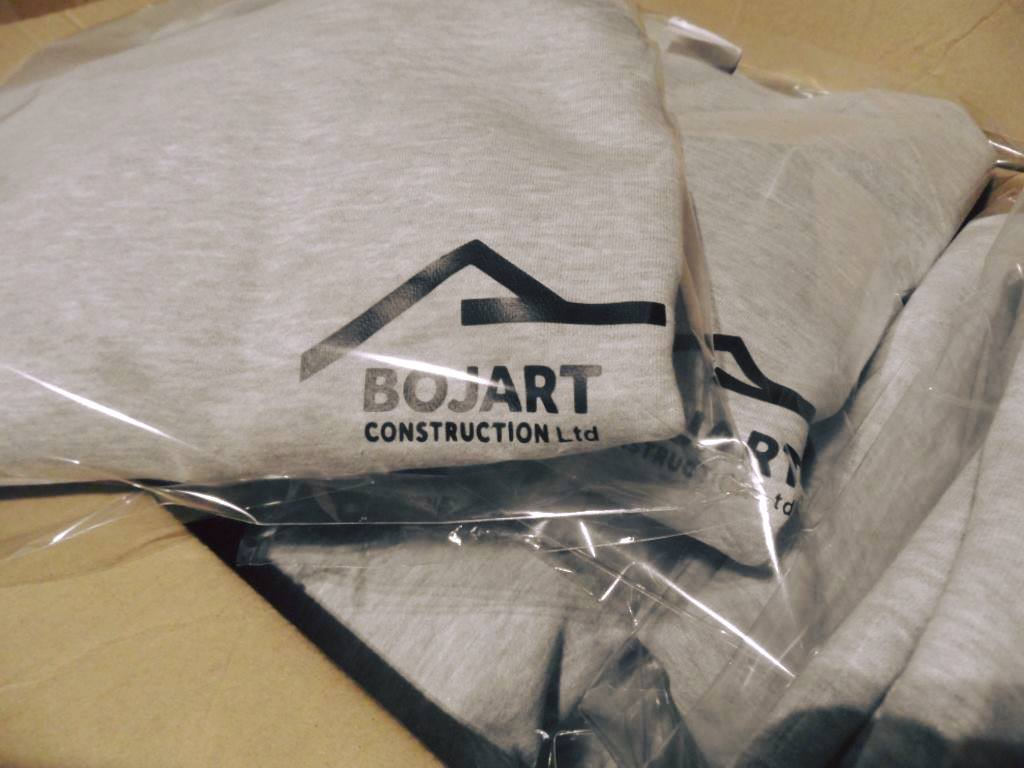 Stos szarych bluz w foliowych opakowaniach z czarnym nadrukiem logo firmy 'BOJART CONSTRUCTION Ltd' z motywem dachu domu.