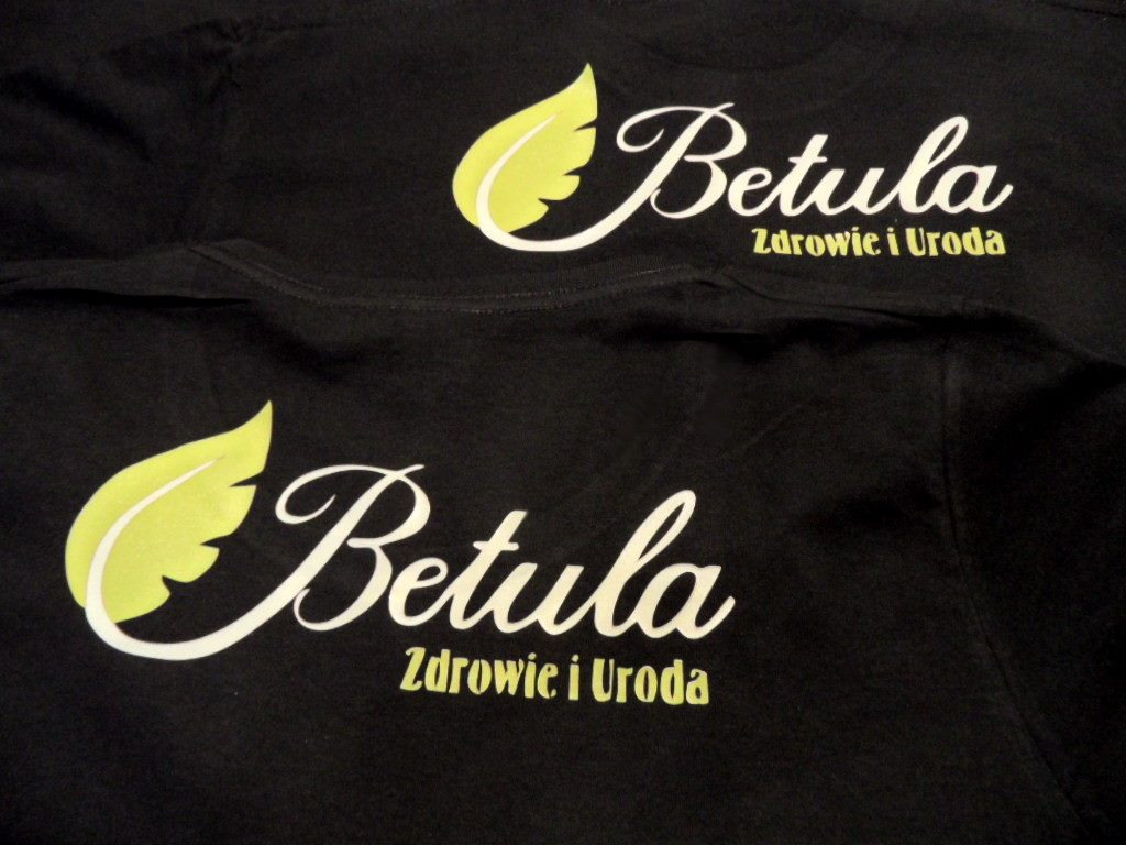 Dwa czarne T-shirty z nadrukowanym logo 'Betula Zdrowie i Uroda' w kolorze białym i żółto-zielonym, ułożone jeden na drugim, widoczne z bliska.