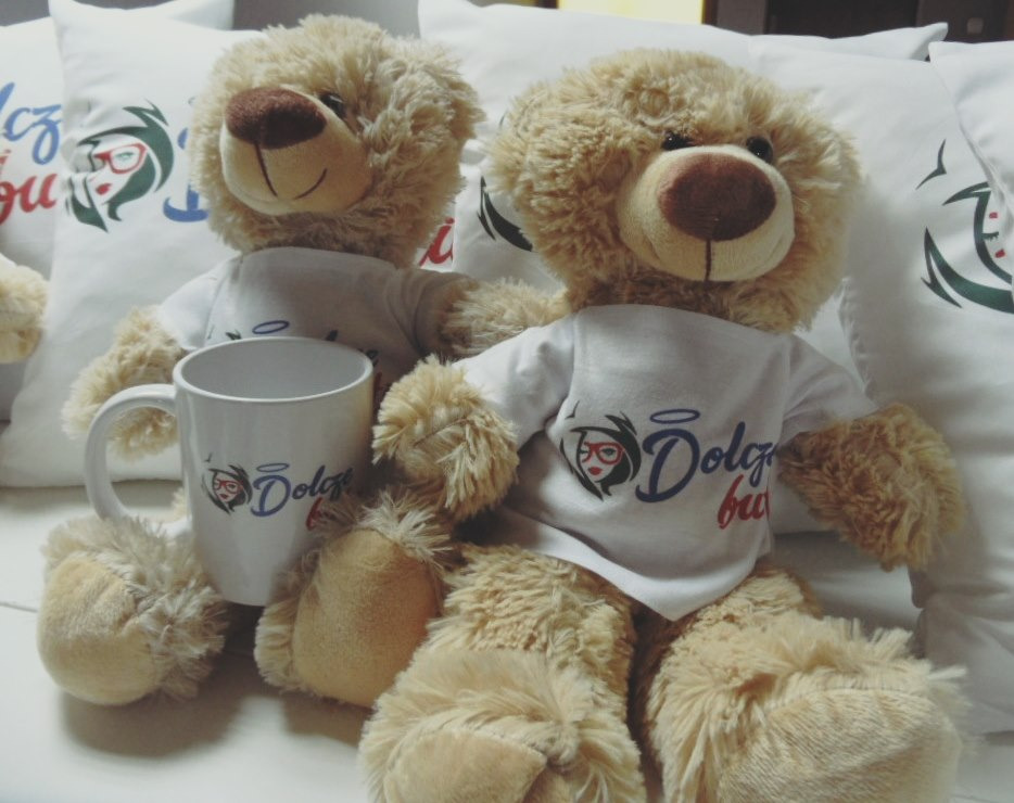 Dwa pluszowe misie w białych koszulkach z nadrukiem, obok biały kubek z tym samym nadrukiem, na tle białych poduszek z nadrukiem.