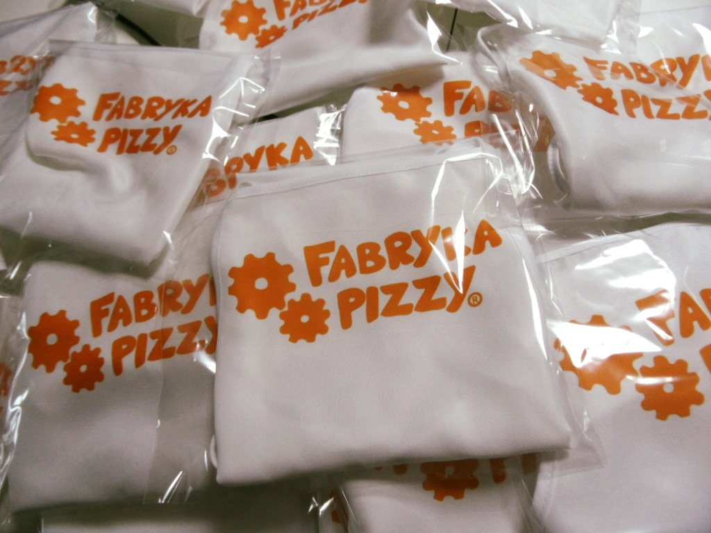 Stos zapakowanych w folię białych fartuchów z pomarańczowym nadrukiem 'Fabryka Pizzy', widoczne detale szwów i zagięć materiału.
