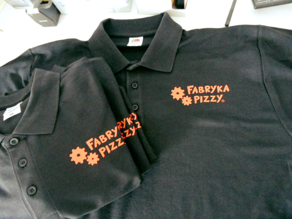 Dwie czarne koszulki polo z pomarańczowym nadrukiem 'Fabryka Pizzy' i graficznymi zębatkami, leżące na białym tle.