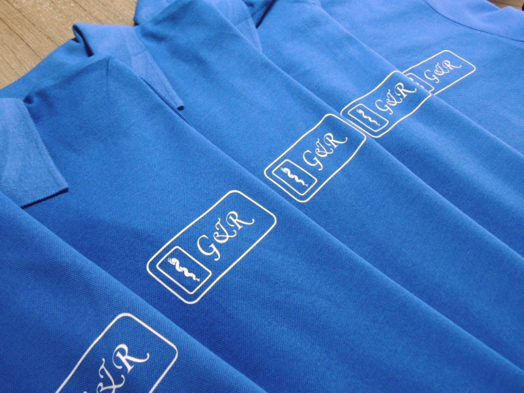 Kilka identycznych, niebieskich koszulek polo z białym nadrukiem logo 'G&R' w prostokątnej ramce, ułożonych ukośnie.