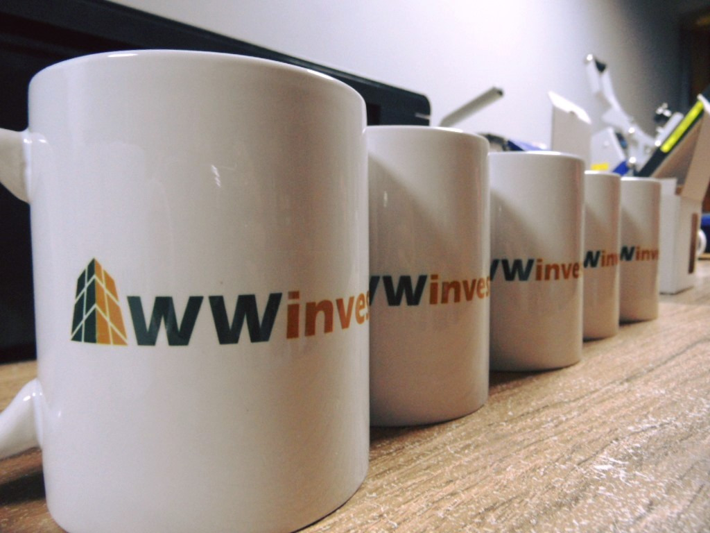 Rząd białych kubków z nadrukowanym logo firmy 'WWinvest' ustawiony na drewnianym blacie, z maszyną do nadruków w tle.