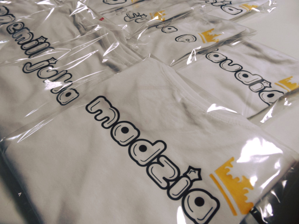 Białe koszulki z czarnym, zaokrąglonym nadrukiem tekstu i żółtą koroną, zapakowane w folię.