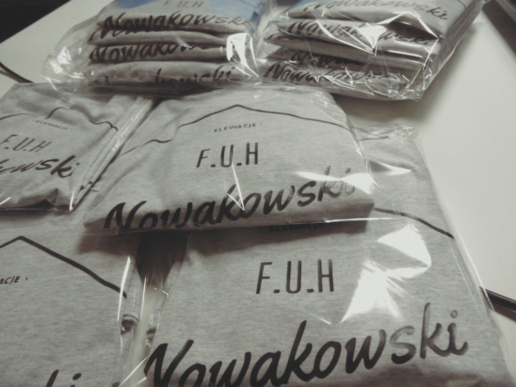 Stos zapakowanych w folię szarych koszulek z czarnym nadrukiem 'F.U.H Nowakowski ELEWACJE' ułożonych na białym tle.