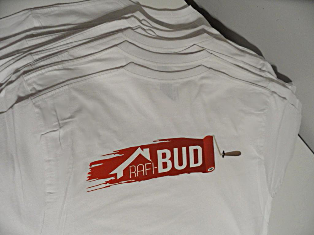 Stos białych koszulek z czerwonym nadrukiem logo firmy budowlanej w formie pędzla malarskiego z napisem RAFI-BUD.