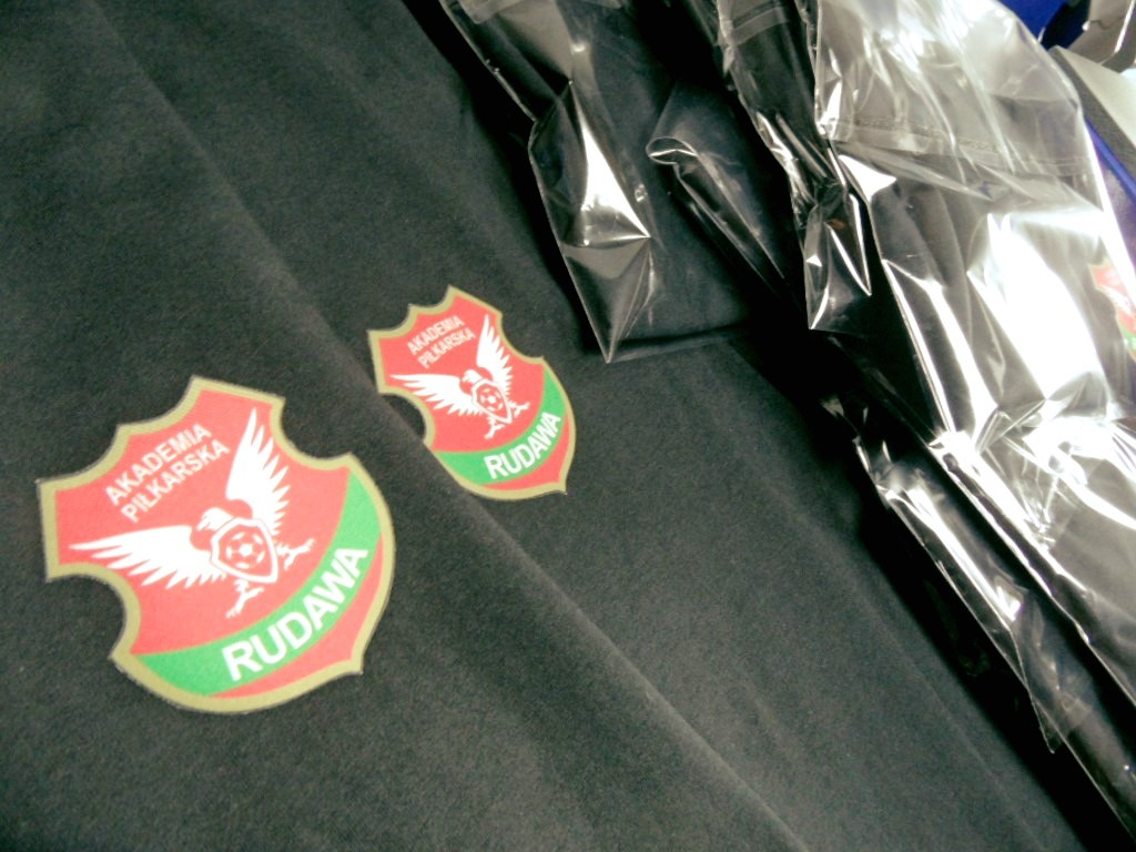 Ciemnoszara bluza dresowa z naszytymi herbami Akademii Piłkarskiej Rudawa, częściowo zapakowana w foliowe opakowanie.