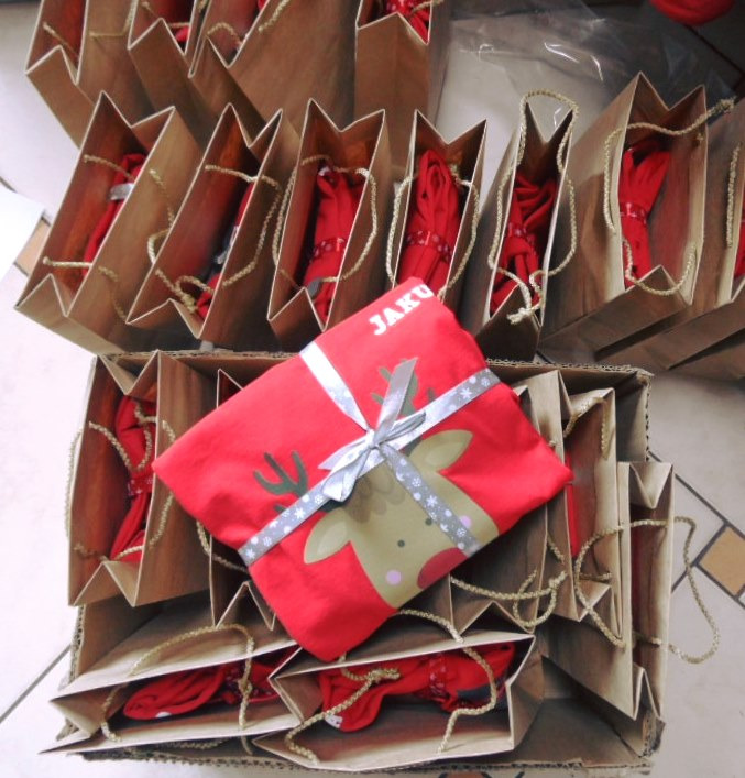 Stos czerwonych koszulek z motywem renifera, zapakowanych w brązowe papierowe torby, jedna koszulka przewiązana srebrną wstążką z imieniem 'JAKU'.