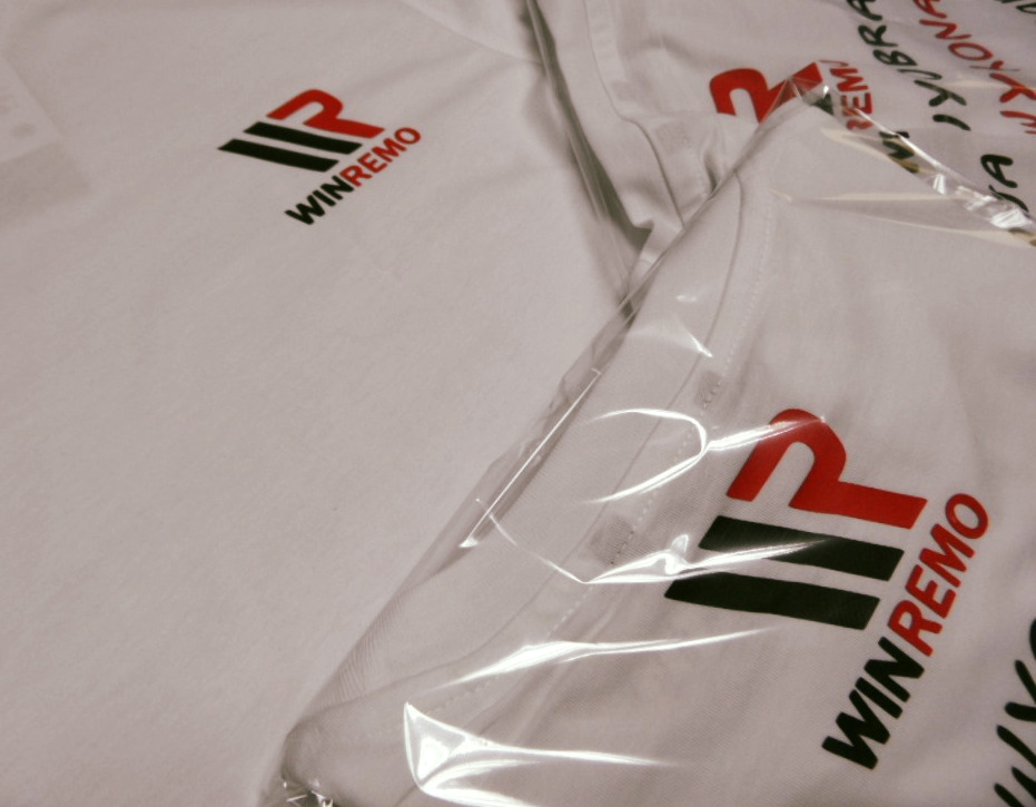 Białe koszulki z nadrukowanym logo WINREMO, część z nich zapakowana w foliowe opakowania.