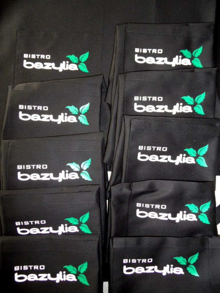Partia czarnych fartuchów roboczych z nadrukowanym logo 'Bistro Bazylia' w kolorze białym i zielonym.