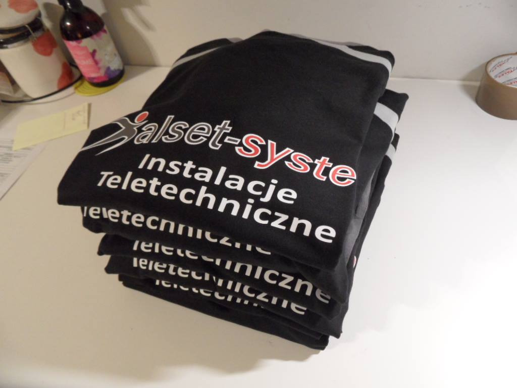 Stos czarnych koszulek z białym i czerwonym nadrukiem 'Salset-System Instalacje Teletechniczne', leżący na białym blacie w otoczeniu kubków i butelki.