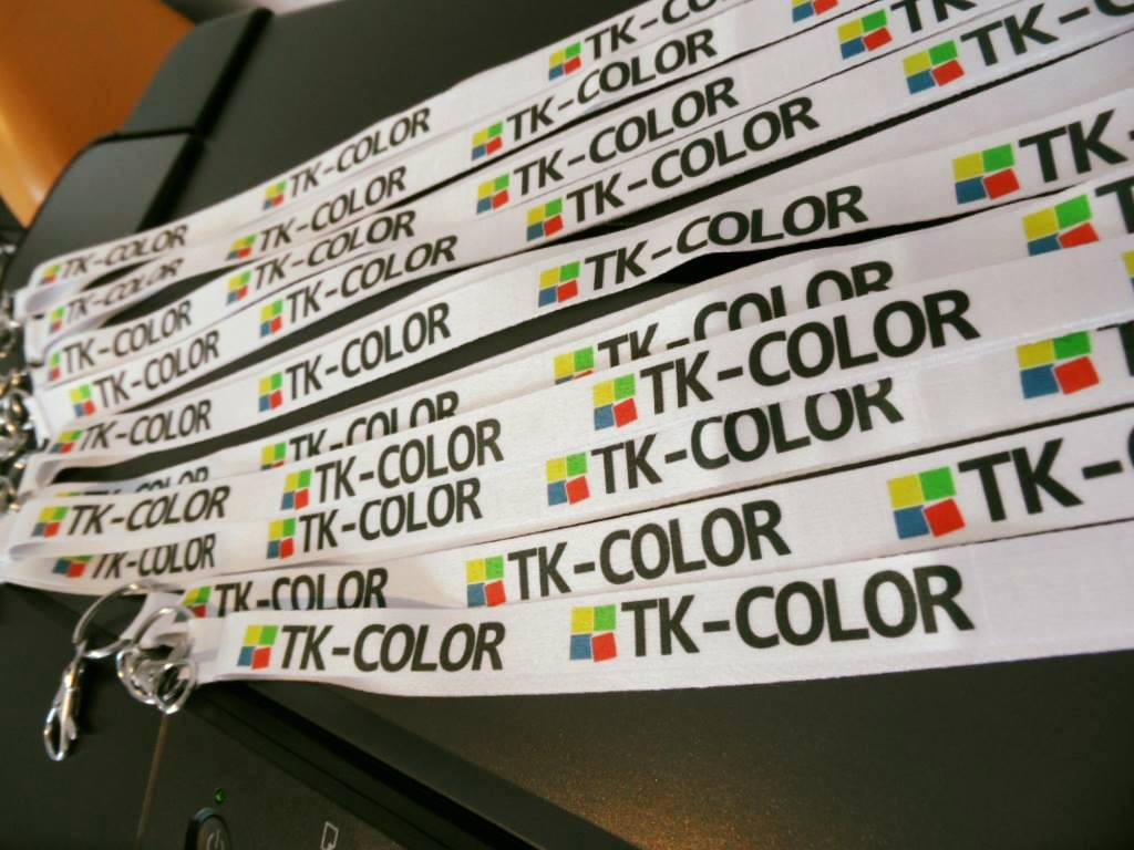 Partia białych smyczy z nadrukowanym czarnym napisem 'TK-COLOR' i kolorowym logo z czterech kwadratów (czerwony, żółty, zielony, niebieski), leżących na czarnej drukarce.