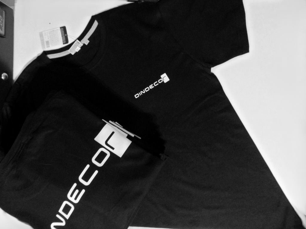 Dwa czarne T-shirty z białym nadrukiem 'DINDECO' leżące na białym tle, jeden złożony, drugi rozłożony, w czarno-białej tonacji.
