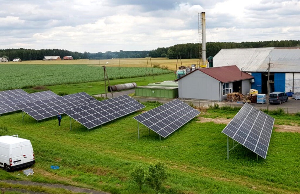 Panel słoneczny ustawiony na łące w pobliżu budynku gospodarczego i pola uprawnego w tle. Widoczny biały samochód dostawczy i osoba w oddali przy panelach.