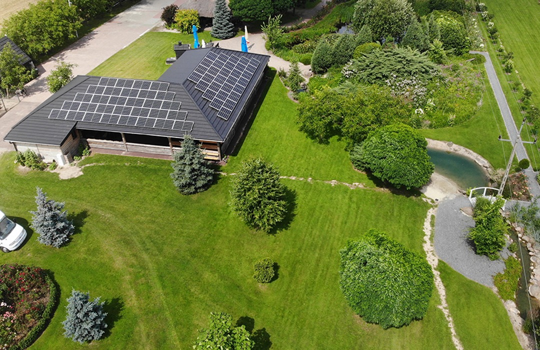 Widok z drona na dom z panelami słonecznymi na dachu, otoczony rozległym, zadbanym ogrodem z oczkiem wodnym i ścieżkami.
