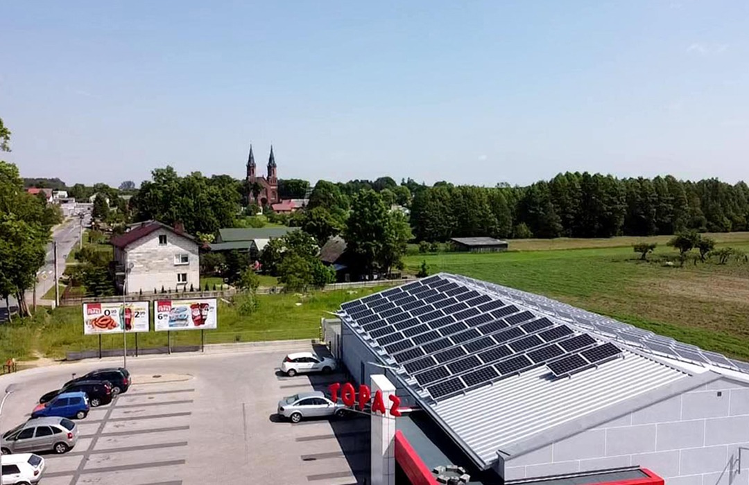 Widok z góry na dach budynku z zainstalowanymi panelami słonecznymi, na parkingu zaparkowane samochody, w tle zabudowania wiejskie i kościół z dwiema wieżami.