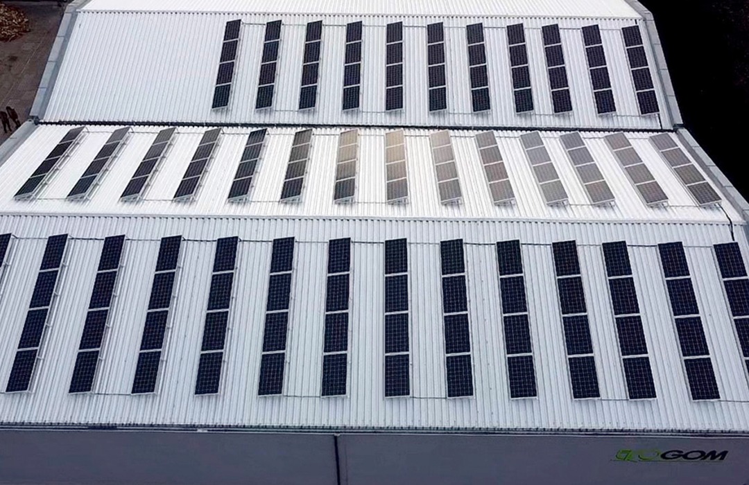 Rząd paneli fotowoltaicznych na falistym, białym dachu przemysłowym, widok z góry.