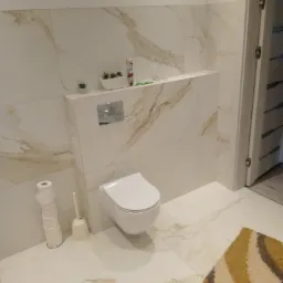 Nowoczesna toaleta podwieszana w łazience wykończonej płytkami imitującymi marmur. Widoczne akcesoria łazienkowe i fragment dywanika w neutralnych barwach.