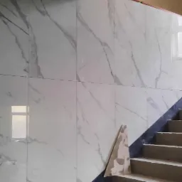 Ściana schodów wyłożona białymi płytkami imitującymi marmur, widoczny surowy stan schodów przed wykończeniem, worek z zaprawą na górze schodów.