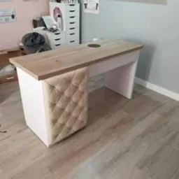 Jasny blat drewniany biurka z otworem na kable, wsparty na białych nogach, z jedną nogą obitą beżową pikowaną tkaniną. W tle widoczny regał z szufladami i różowa ściana.