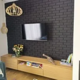 Ciemna ściana z cegły za telewizorem, pod nią długa, jasna szafka RTV z drewna z dekoracjami i kwiatami, wisząca lampa z drewna w tle.