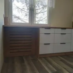Zabudowa okna z grzejnikiem i komodą z białymi frontami szuflad oraz blatem w kolorze drewna, na podłodze panele imitujące drewno.