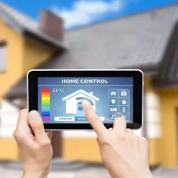 Dłoń dotyka ekranu tabletu z aplikacją 'Home Control' na tle dwukondygnacyjnego domu z garażem i brązową bramą.