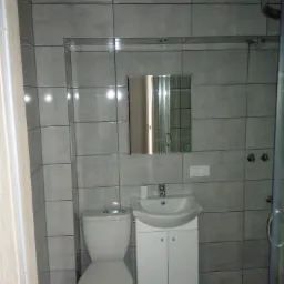 Szara łazienka z toaletą, białą szafką pod umywalką i lustrem z oświetleniem, widok na kabinę prysznicową, płytki ceramiczne na ścianach i podłodze.