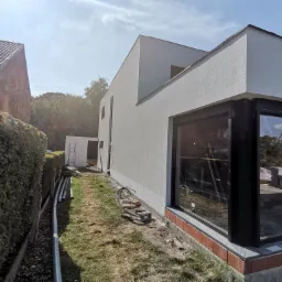 Nowoczesny dom po nałożeniu gładzi zewnętrznej, widoczne okna z czarnymi ramami, ceglana podmurówka, rury i drabina w tle.