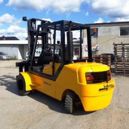 Żółty wózek widłowy Balkancar sfotografowany z tyłu na betonowym placu, z paletami i budynkiem w tle.