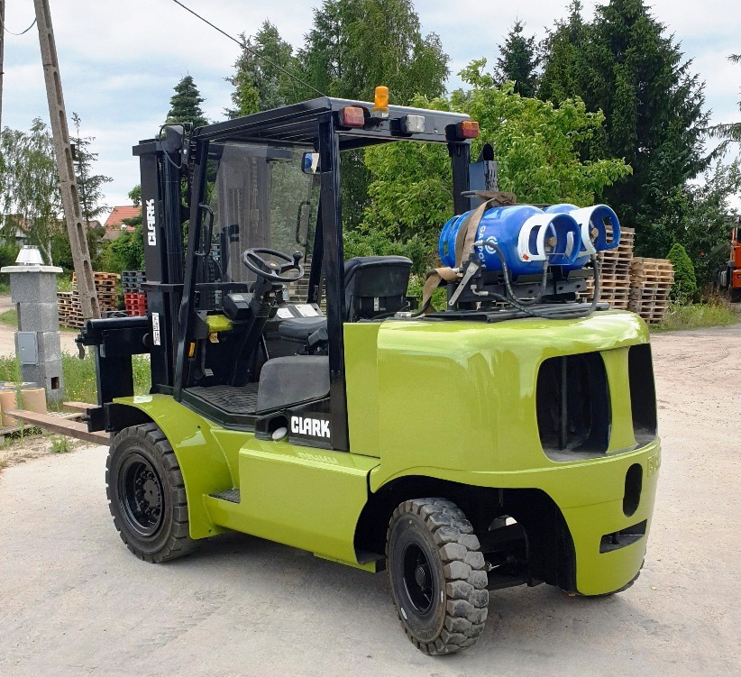 Nowy wózek widłowy marki Clark w kolorze oliwkowym, z zamontowanymi dwoma niebieskimi butlami gazowymi na tylnej części, stojący na betonowym podłożu na zewnątrz.