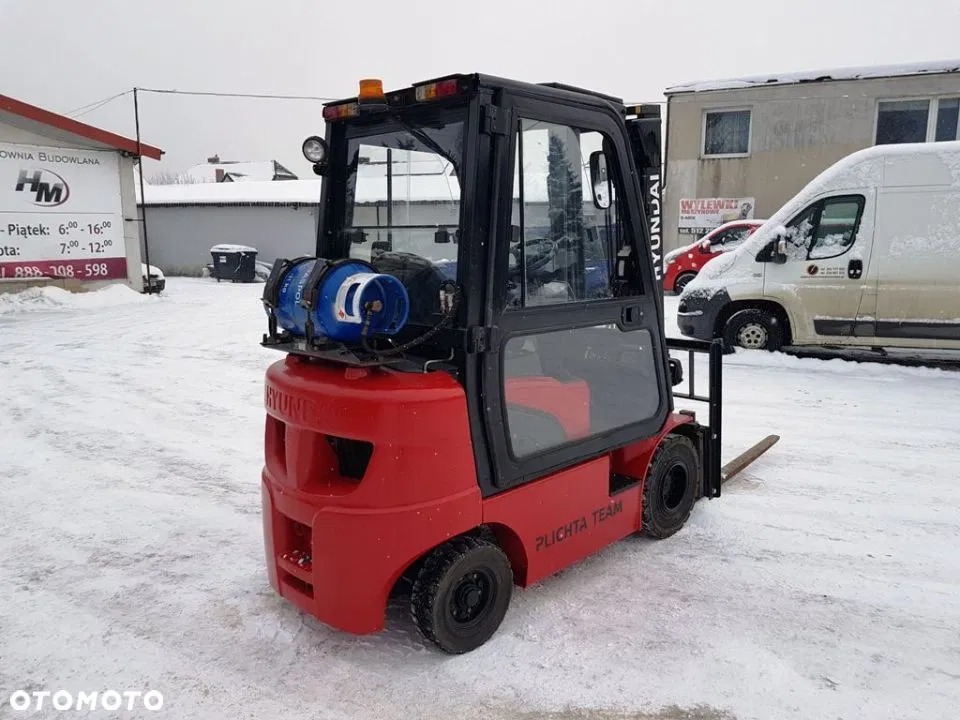 Czerwony wózek widłowy Hyundai z butlą gazową na zewnątrz, stojący na śniegu przed budynkiem z szyldem 'Budownia Budowlana' i zaparkowanym białym vanem.