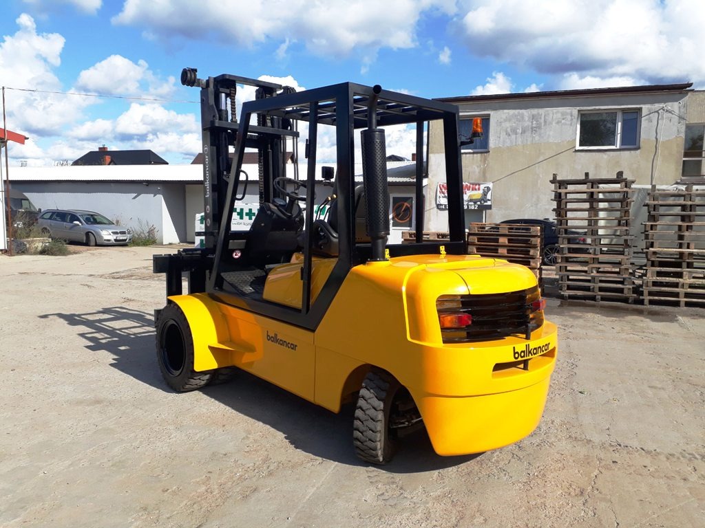 Żółty wózek widłowy Balkancar sfotografowany z tyłu na betonowym placu, z paletami i budynkiem w tle.