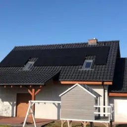 Dom jednorodzinny z zainstalowanymi czarnymi panelami słonecznymi na dachu z dachówki w kolorze grafitowym, widoczne okna dachowe, mały drewniany domek dla dzieci na trawniku.