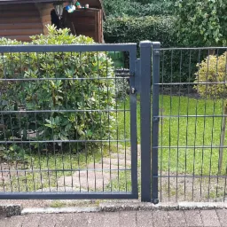 Otwarta, nowoczesna furtka z metalowej siatki ogrodzeniowej na tle zadbanego ogrodu z krzewami i ścieżką.