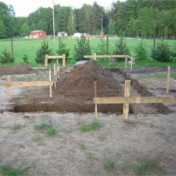 Wytyczanie fundament&oacute;w budynku z użyciem drewnianych szalunk&oacute;w i wykopanych row&oacute;w, z hałdą ziemi pośrodku, w tle zielona łąka i zabudowania gospodarcze.