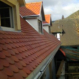 Krol-DACH - Dach pokryty czerwoną dachówką karpiówką, z trzema lukarnami, widoczne orynnowanie i fragmenty konstrukcji drewnianej, w tle inne budynki i zieleń.