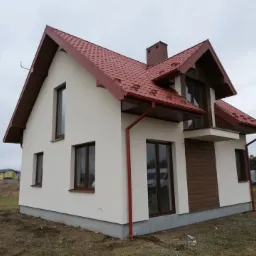 Nowoczesny dom jednorodzinny z beżową elewacją, brązową stolarką okienną i dachem z czerwonej dachówki, widoczny z poziomu gruntu w pochmurny dzień.