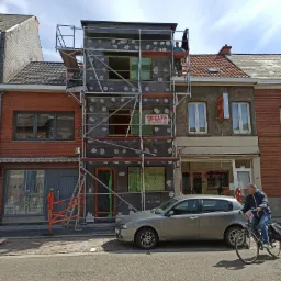 Prace na wysokościach przy budynku w trakcie nakładania izolacji na elewację, widoczne rusztowania, folie okienne, pracownik na rusztowaniu oraz zaparkowany samochód osobowy na ulicy.