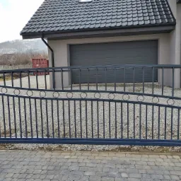 Nowoczesna, metalowa brama wjazdowa w kolorze grafitowym z dekoracyjnymi okręgami, na tle domu z szarą elewacją i garażem, widok z zewnątrz na kostce brukowej.