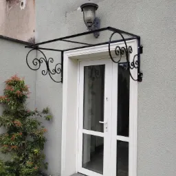 Zadaszenie nad białymi drzwiami wejściowymi z dekoracyjnymi elementami kutymi i lampą ścienną, widok z zewnątrz.