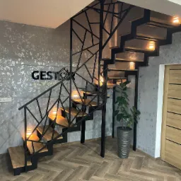 Schody metalowe z drewnianymi stopniami i geometryczną balustradą w Bielawie, oświetlone punktowymi lampkami, obok donica z rośliną, na ścianie logo firmy.