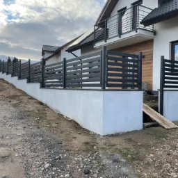 Nowoczesne ogrodzenie panelowe w kolorze antracytowym, zamontowane na podmurówce z białego betonu, w tle dom z balkonem z metalową balustradą.